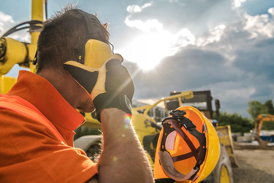 A construction worker takes off a helmet and ear protectors with construction equipment visible in the background